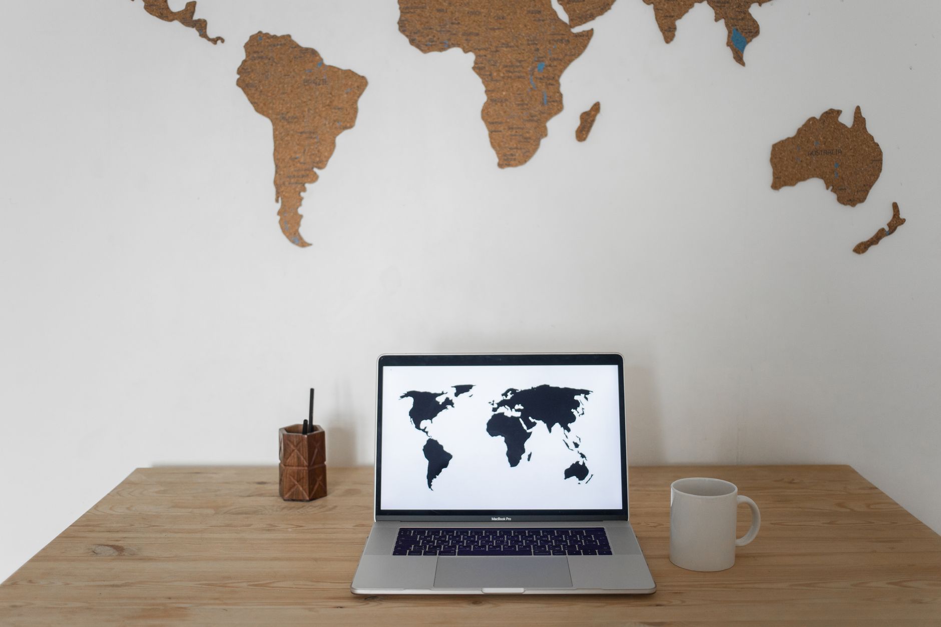 Wooden desk with laptop displaying world map, cork desk organizer, and white mug against wall with cork world map