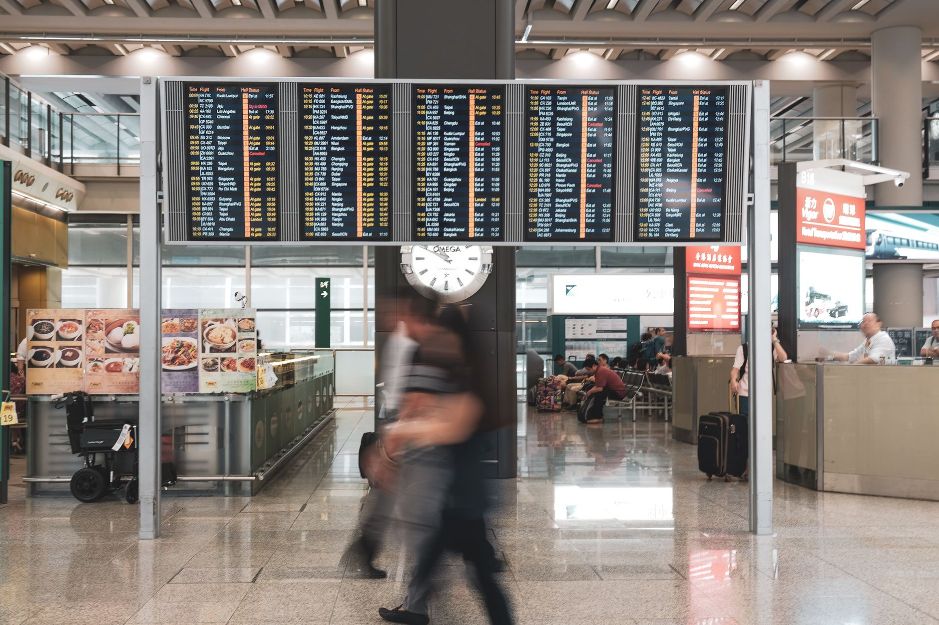 Passengers walking through an airport terminal corridor with a flight information display board overhead
