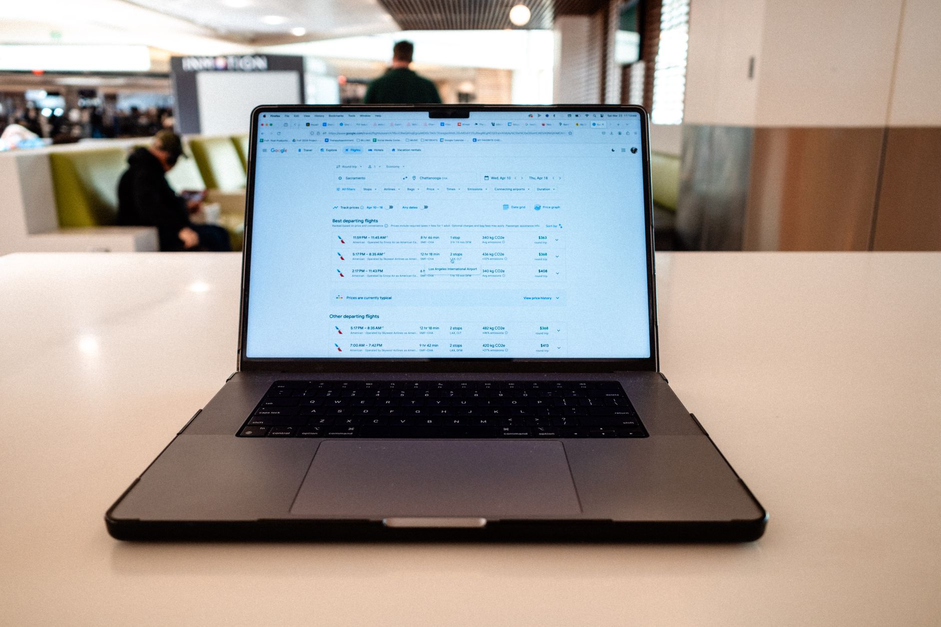 Laptop showing flight search results with multiple departure options and pricing in airport terminal