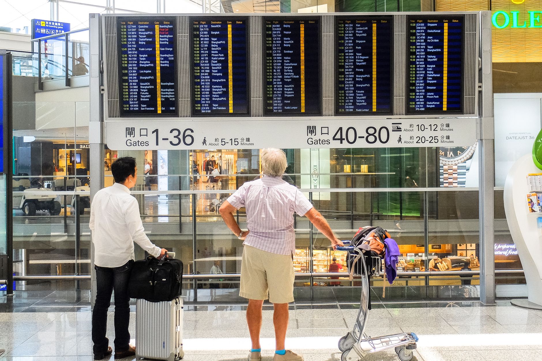 Two travelers checking a departure board at an airport gate, with luggage and a bicycle nearby