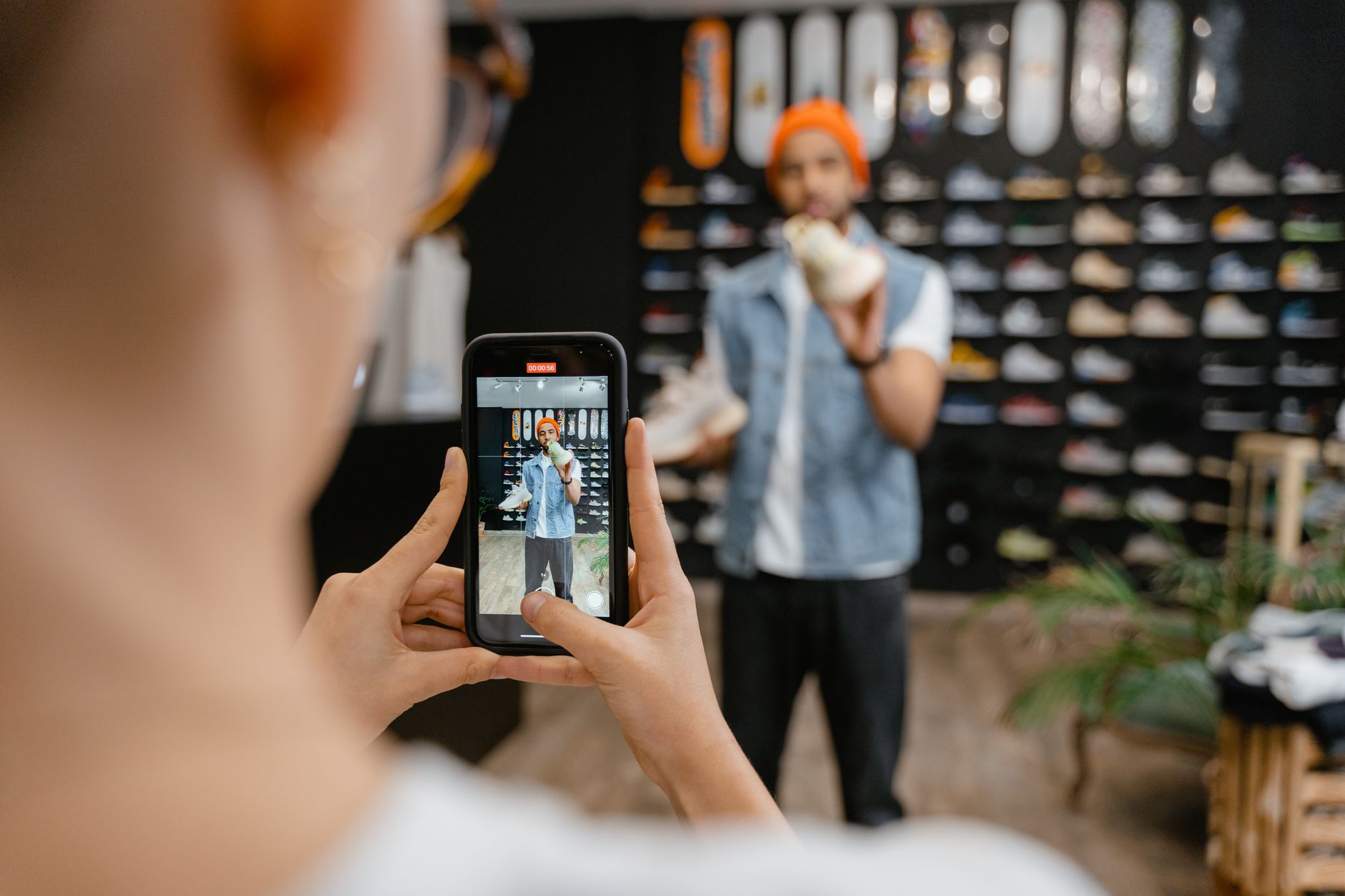 Person holding smartphone recording another person at a sneaker store with shoe wall display in background